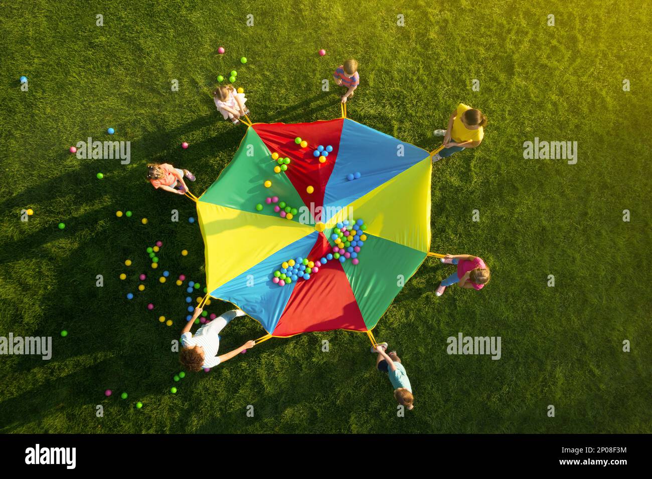Group of children and teachers playing with rainbow playground parachute on green grass, top