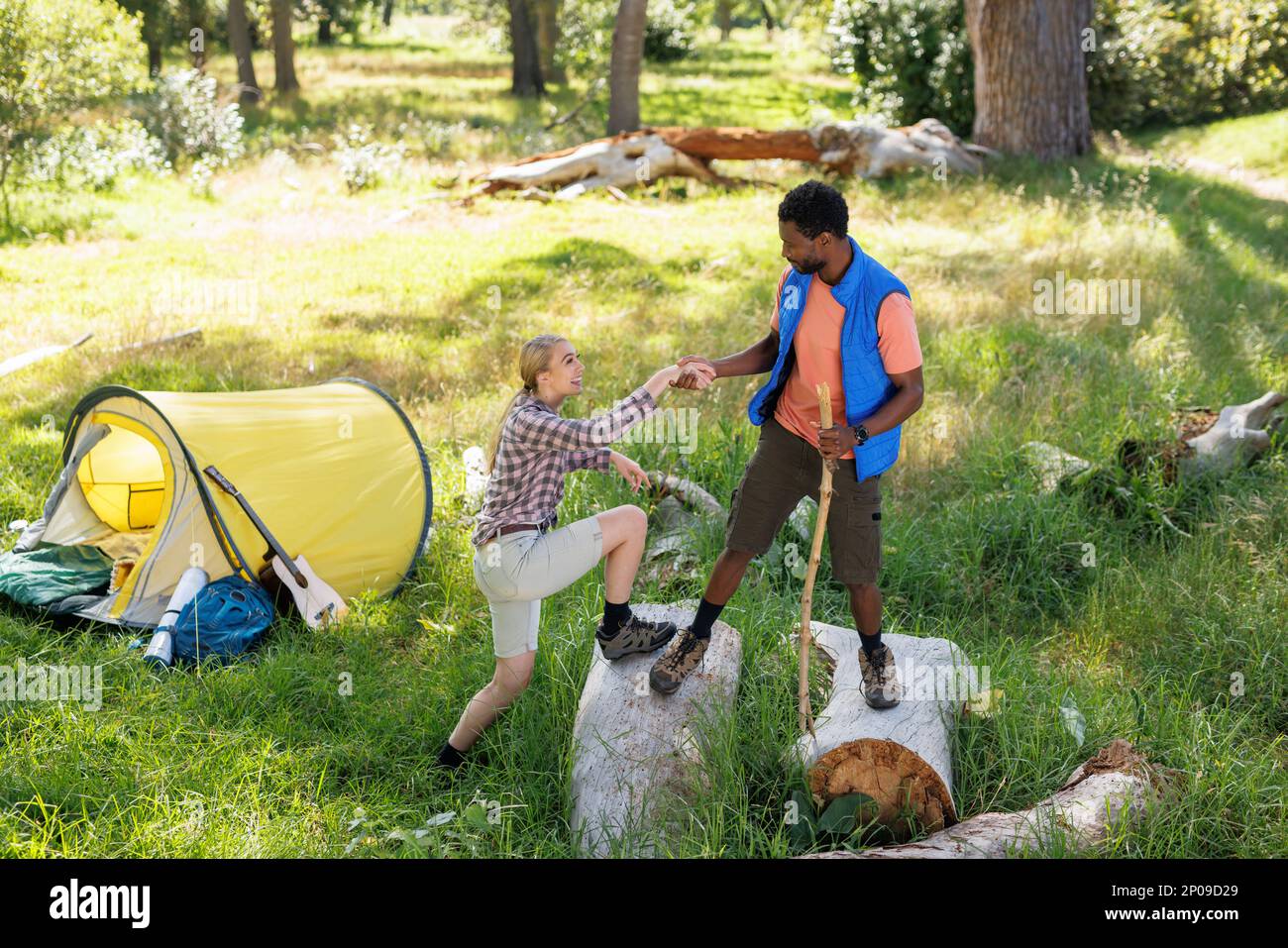Happy, diverse couple on camping holiday climbing on logs by their tent in countryside Stock Photo
