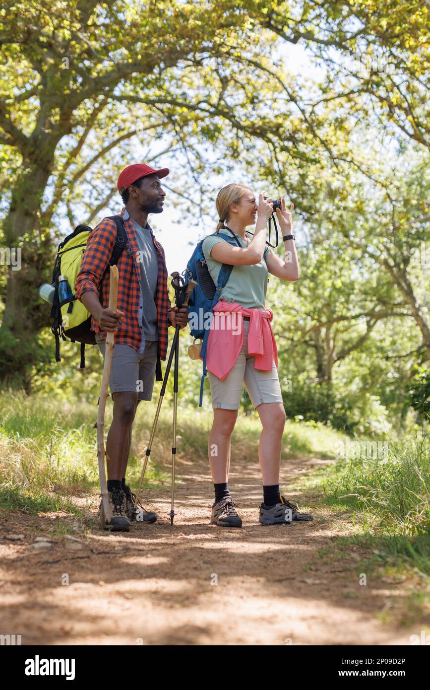Vertical of happy, diverse couple with backpacks hiking through forest, taking photos, copy space Stock Photo