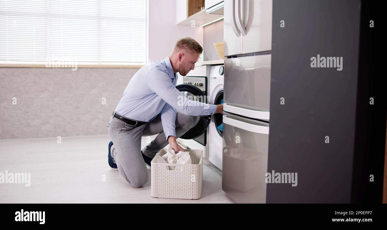Young Man Loading Clothes Into Washing Machine In Kitchen Stock Photo