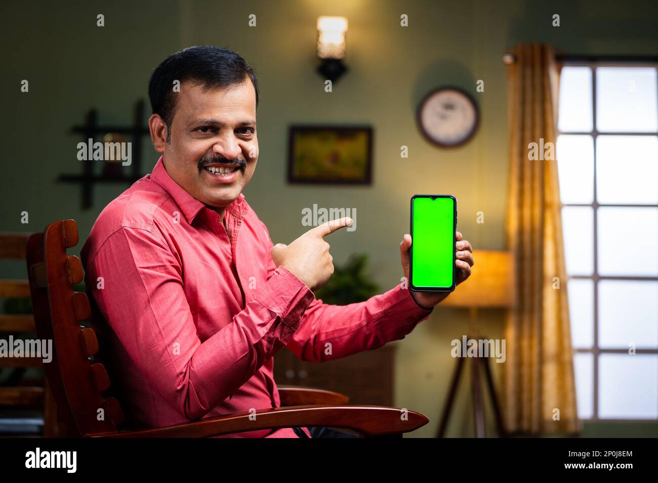 Happy smiling middle aged man showing green screen mobile phone by pointing finger by looking camera at home - concept of app promotion, advertisement Stock Photo