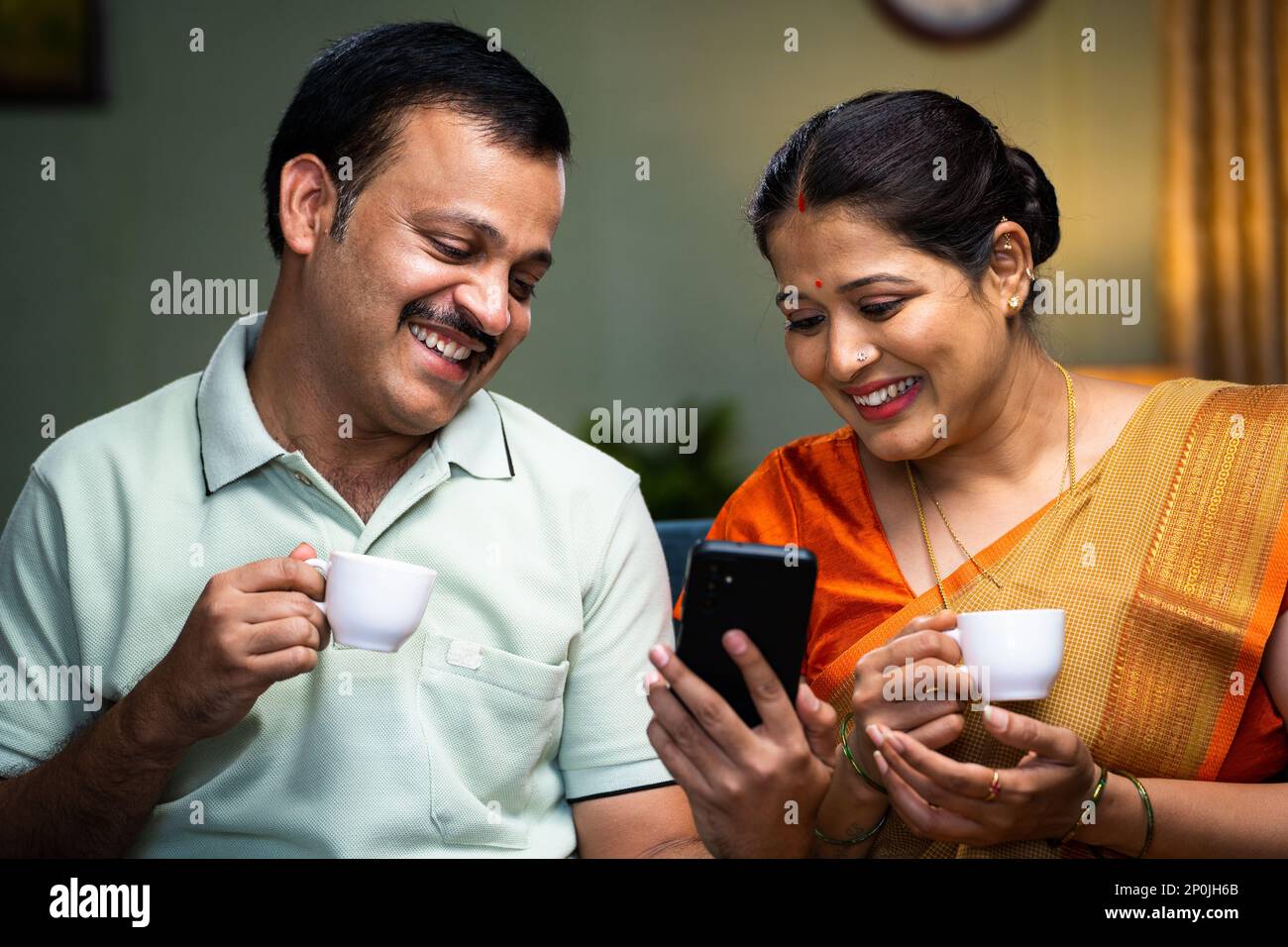 Happy smiling couple drinking tea while watching mobile phone at home - concept of relaxation, refreshment and relationship bonding. Stock Photo