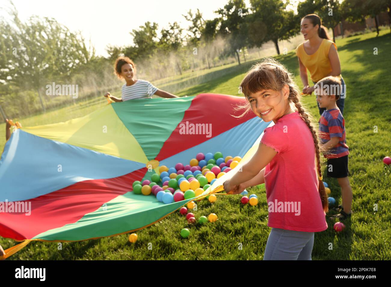 Group of children and teachers playing with rainbow playground parachute on green grass. Summer