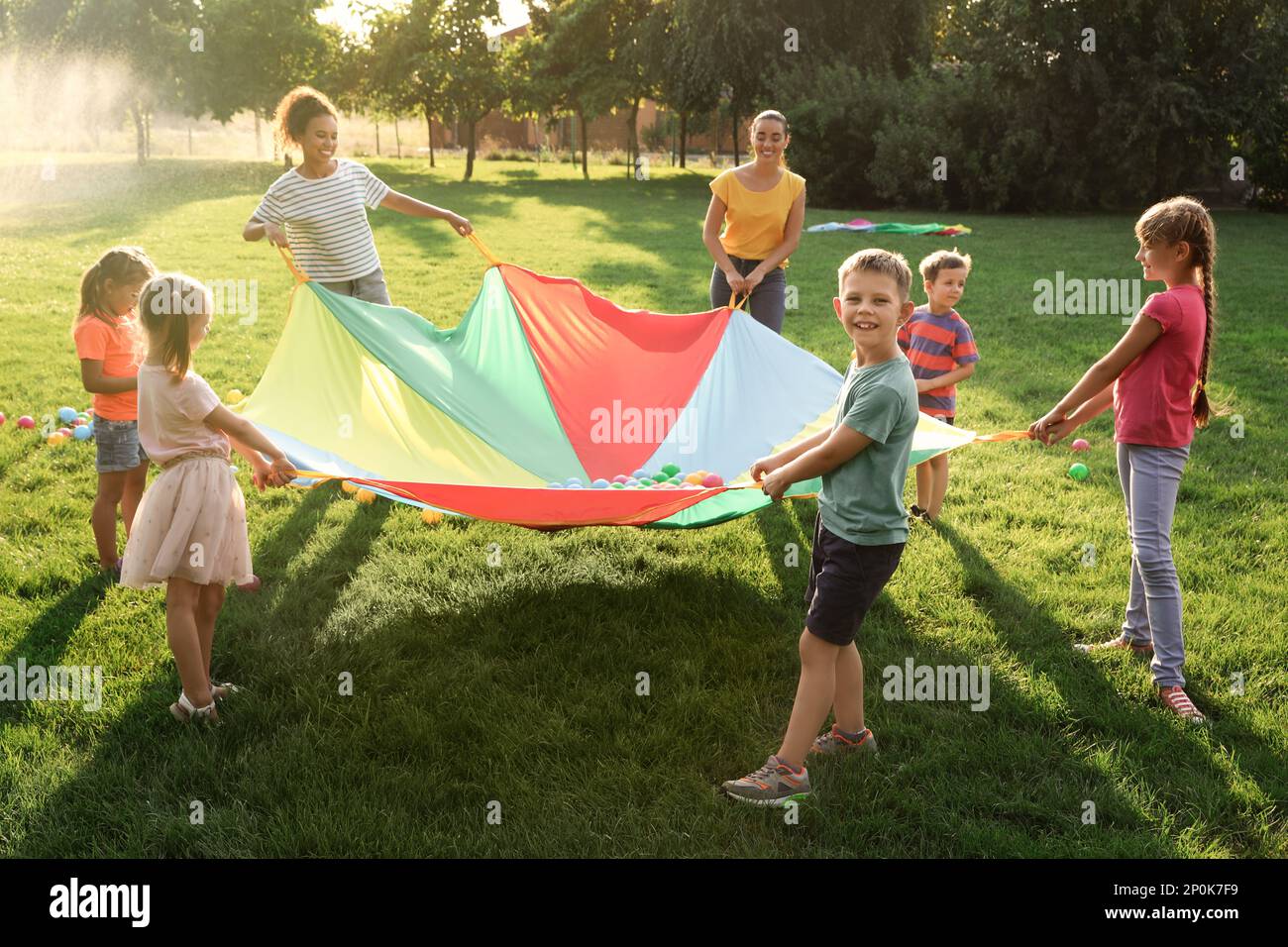 Group of children and teachers playing with rainbow playground parachute on green grass. Summer