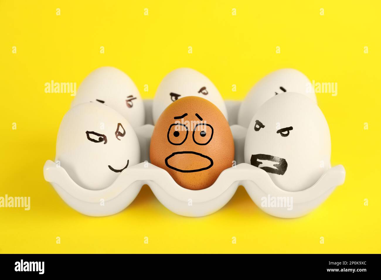 Brown egg with scared face among aggressively disposed white ones in holder on yellow background. Stop racism Stock Photo