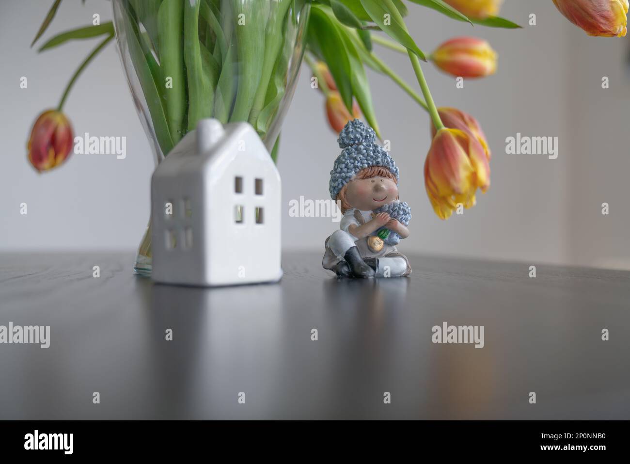 Figure of a girl with flowers and a house near a vase with tulips against the background of a white wall. Stock Photo