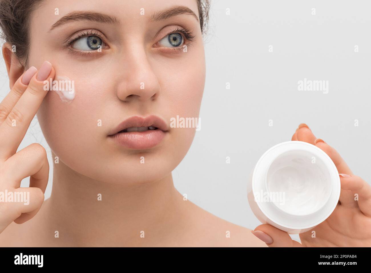 Young beautiful girl applies cream to her face, close-up. Long hair, big blue eyes and clear skin. Stock Photo