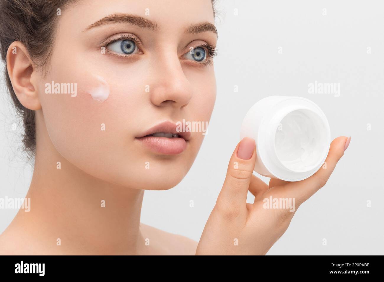 Young beautiful woman applies cream to her face, close-up. Long hair, big blue eyes and clear skin. Stock Photo