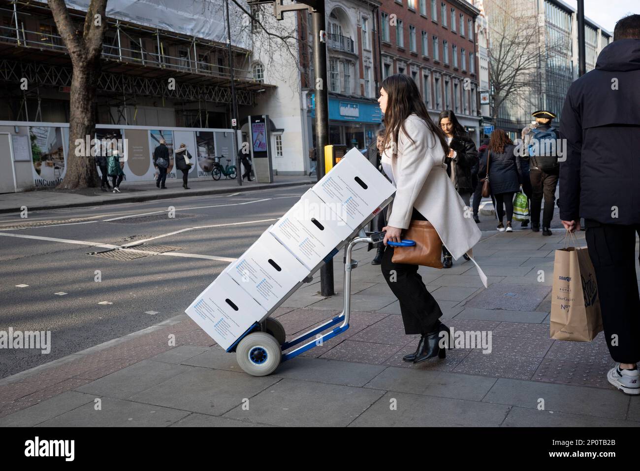 A young employee pushes a load of boxes and uses a trolley along High Holborn, on 2nd March 2023, in London, England. Stock Photo