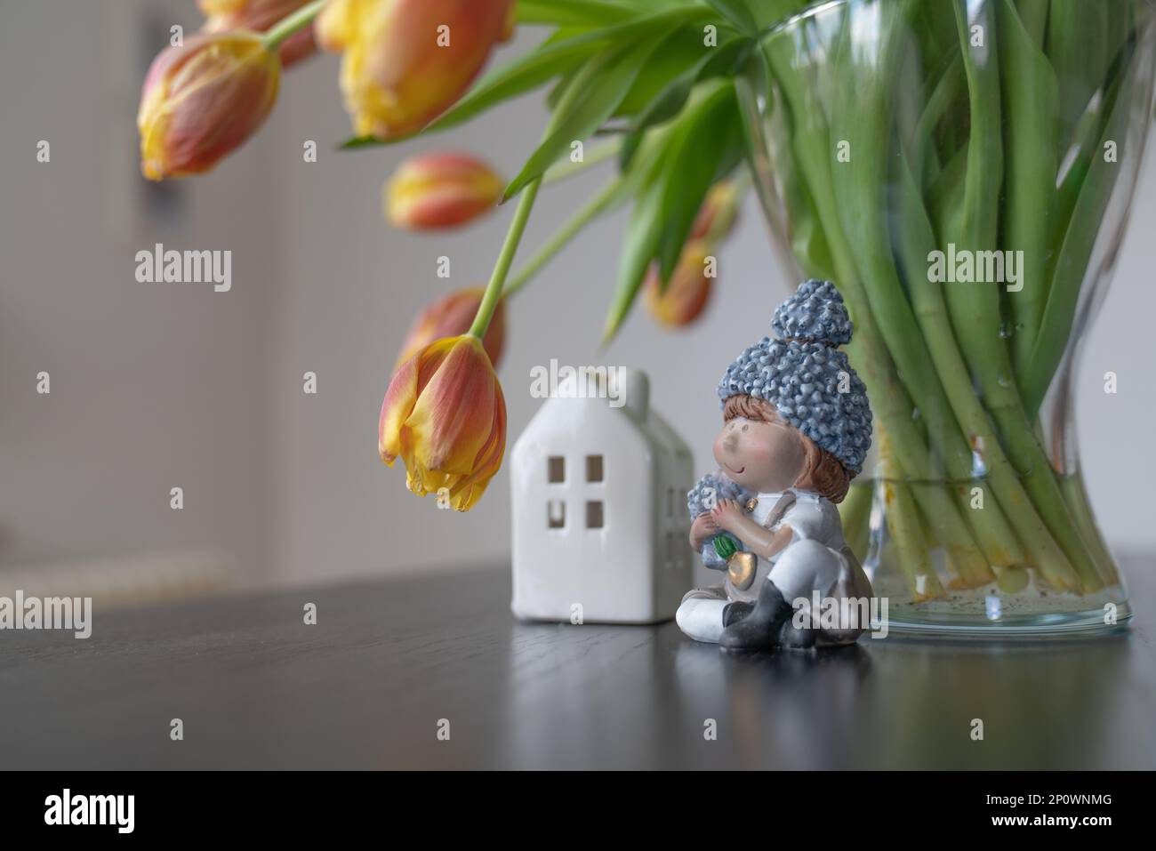Figure of a girl with flowers and a house near a vase with tulips against the background of a white wall. Stock Photo
