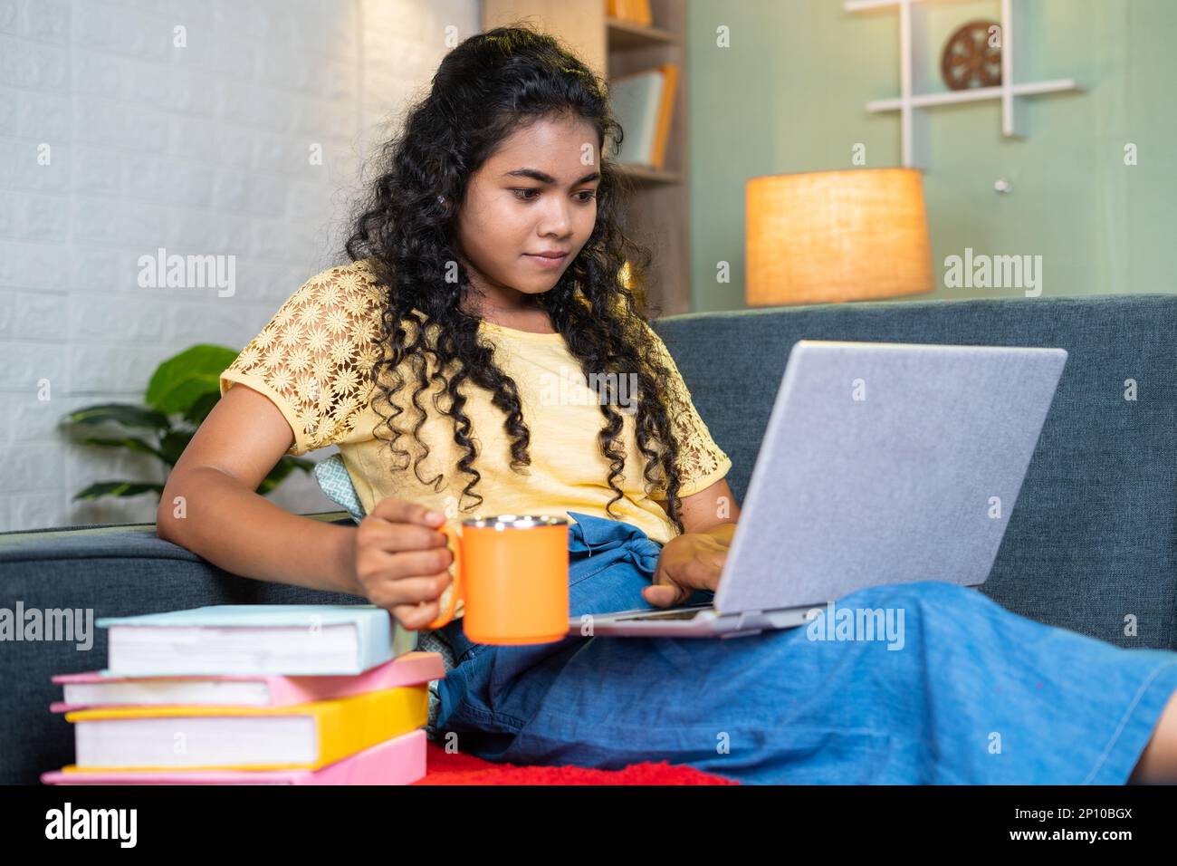 Young indian girl drinking coffee or tea while using laptop during examination while sitting on sofa at home with books - concept of refreshment Stock Photo