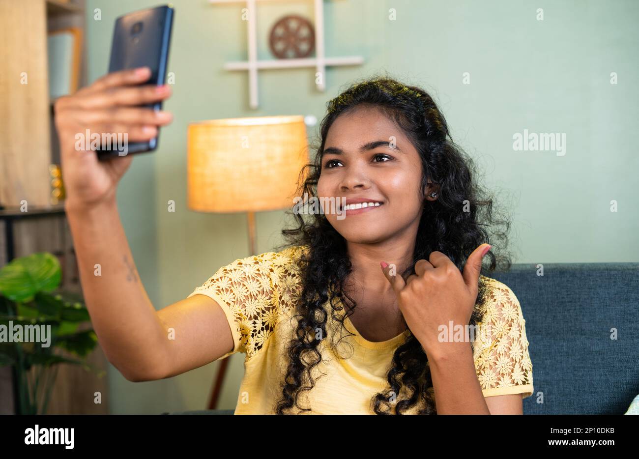 Happy smiling young girl taking selfie on mobile phone by showing hands gesture at home - concept of technology, social media and addiction Stock Photo