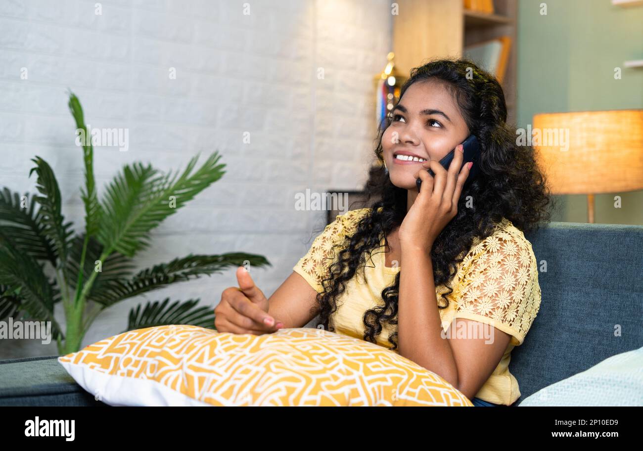Smiling young teenager girl talking on mobile phone with friends while sitting on sofa at home - concept of communication, distance Relationship and Stock Photo