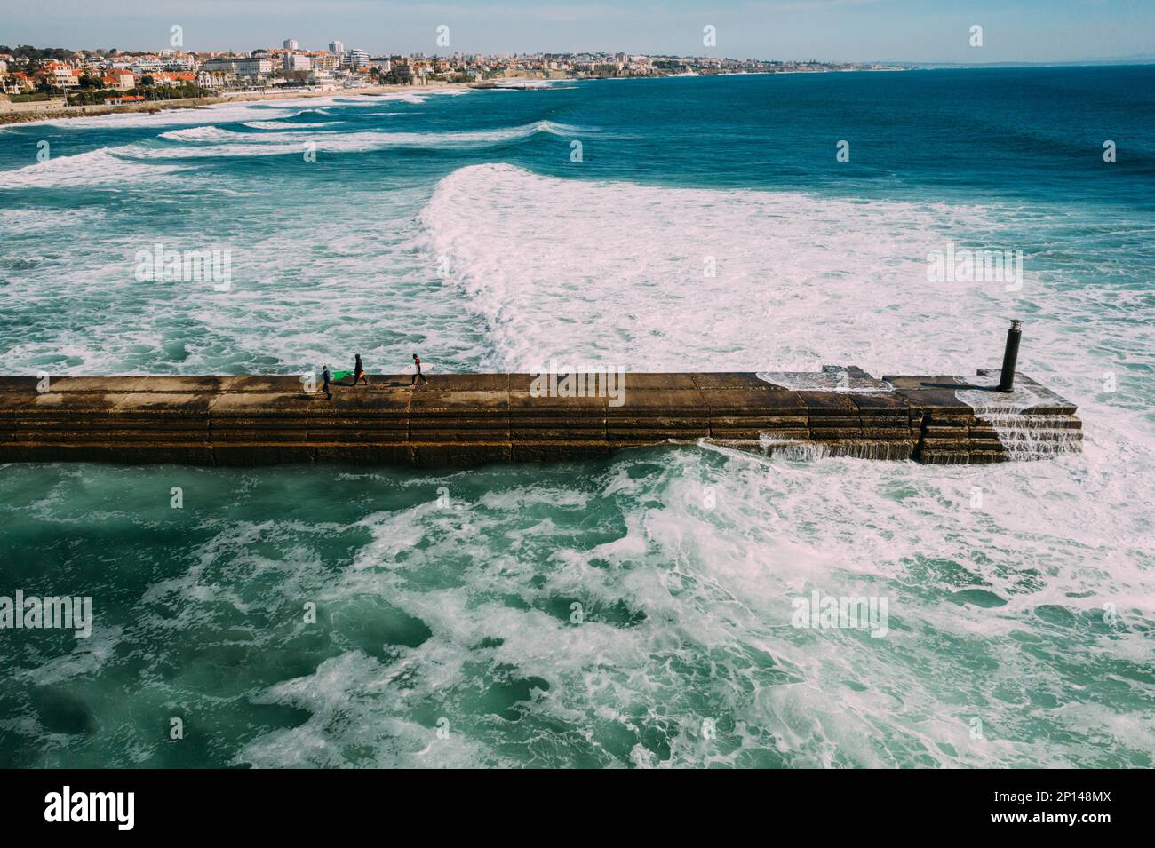 Surfers carrying their boards at a pier in Cascais, Portugal with giant swell visible Stock Photo