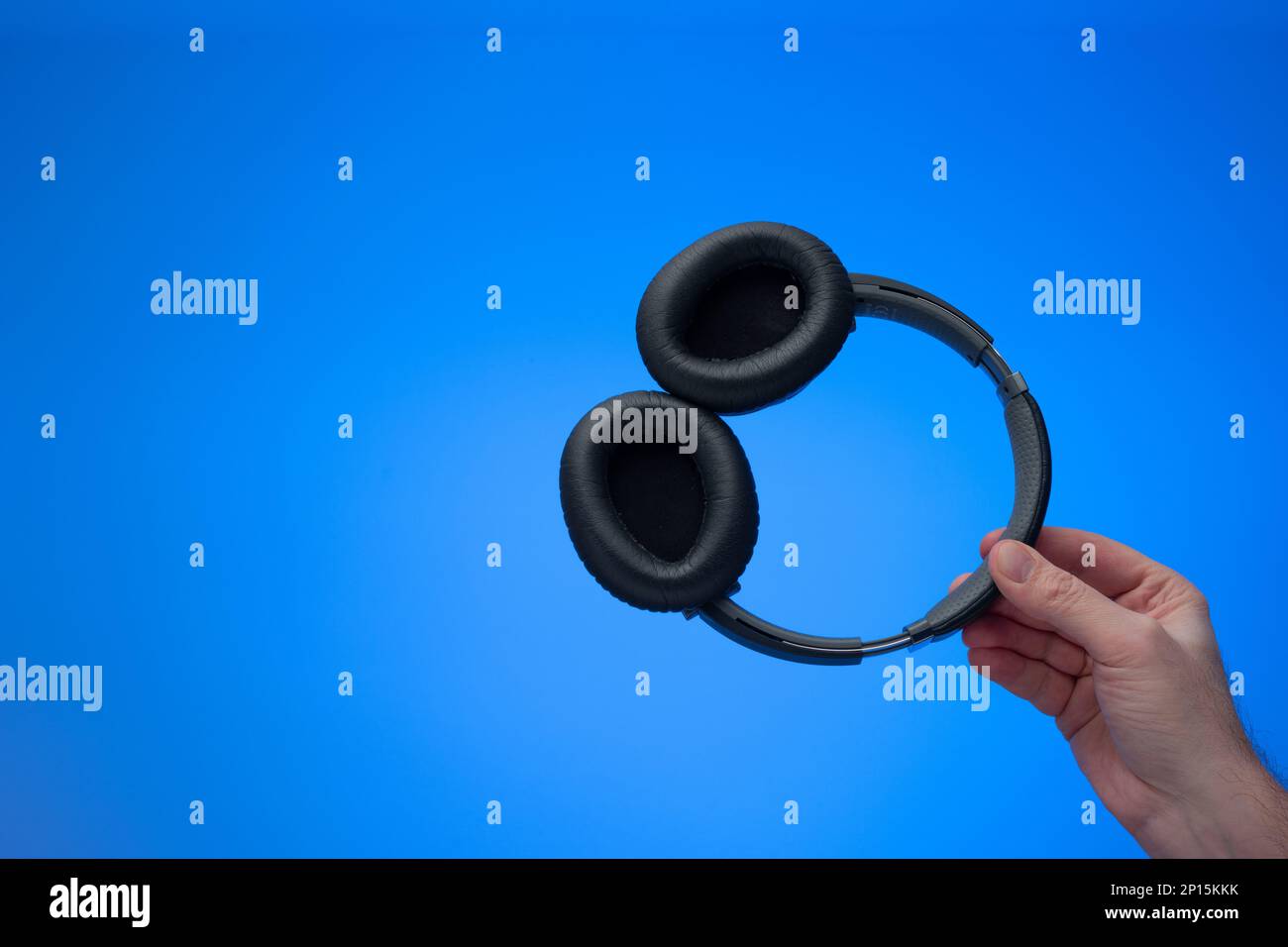 Generic wireless over the ear headphone held in hand by Caucasian male hand. Close up studio shot, isolated on blue background. Stock Photo