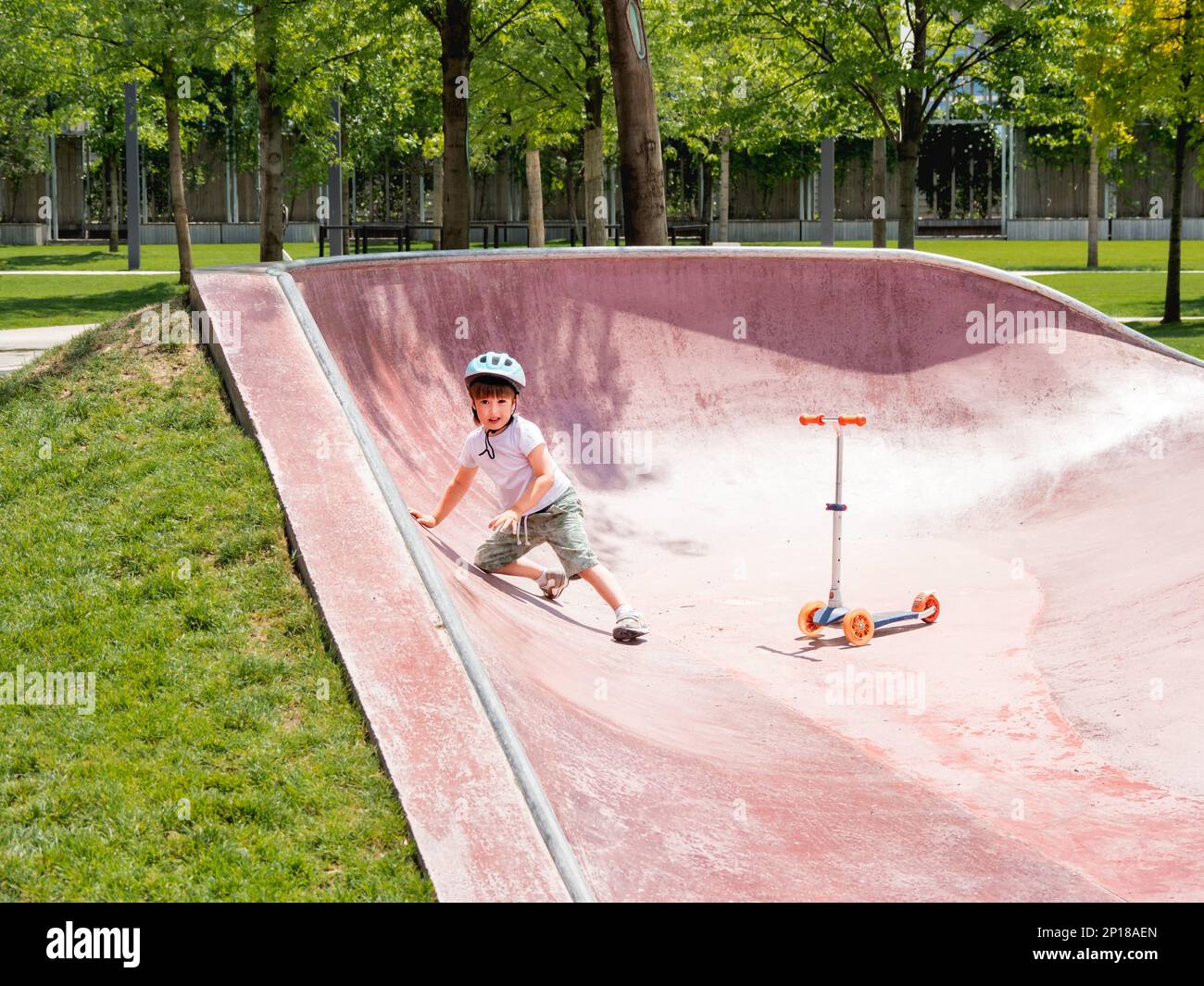 Little boy fell off kick scooter while riding in skate park. Special concrete bowl structures in urban park. Training to skate at summer. Stock Photo