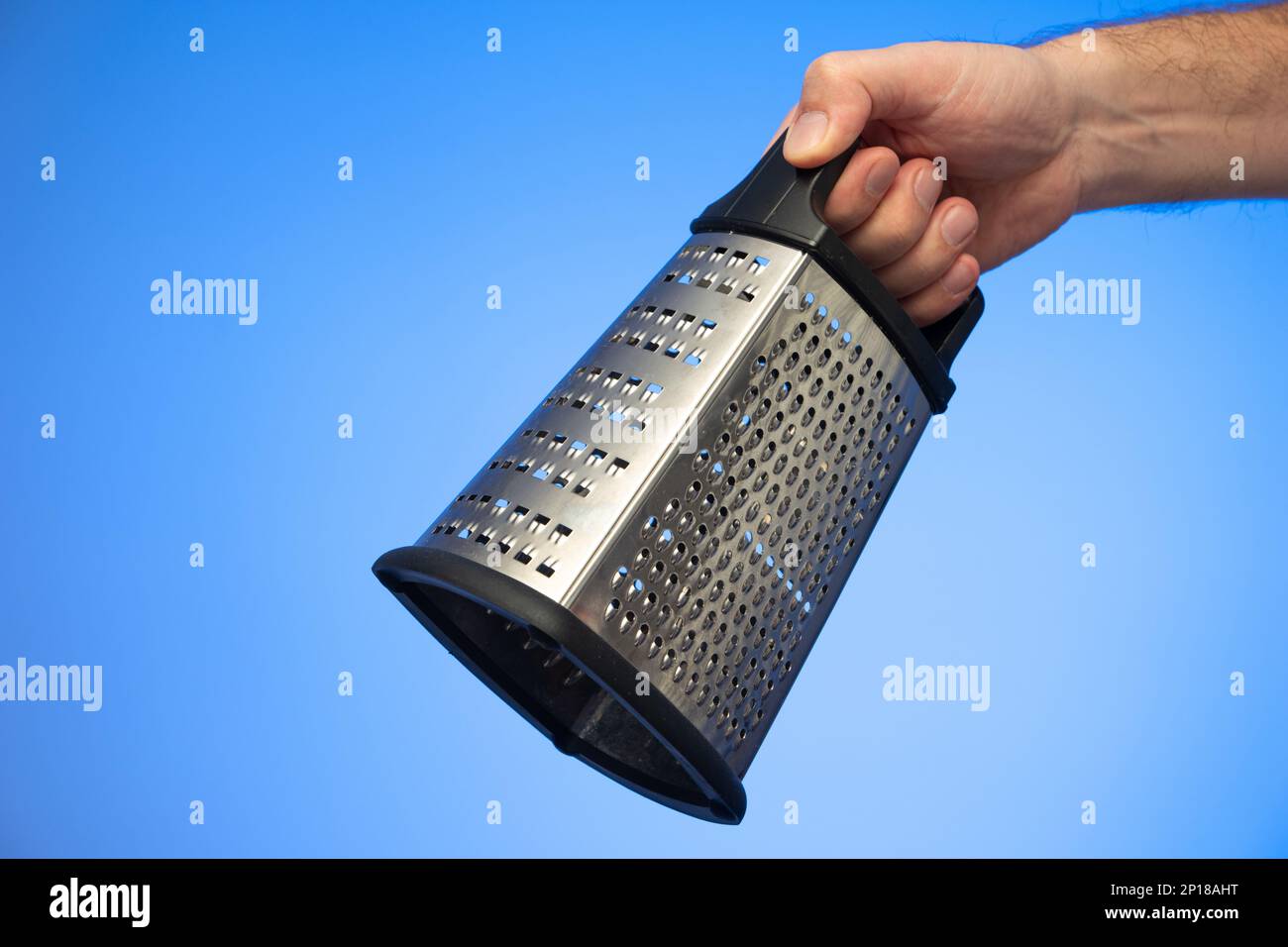 Stainless metal kitchen grater with plastic handle held in hand by Caucasian male hand studio shot isolated on blue background. Stock Photo