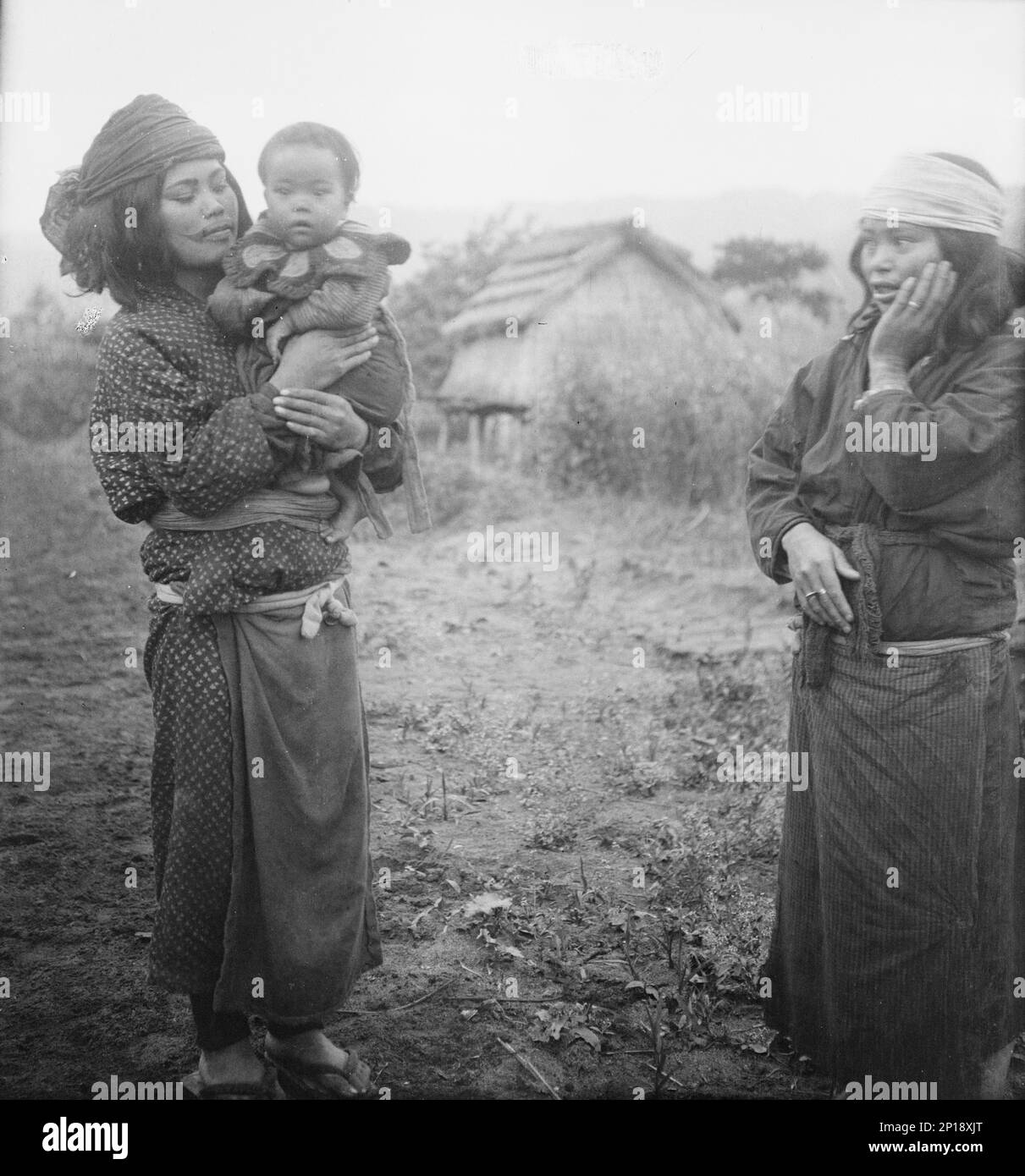 Two Ainu woman, one holding a child, standing outside, 1908. Stock Photo