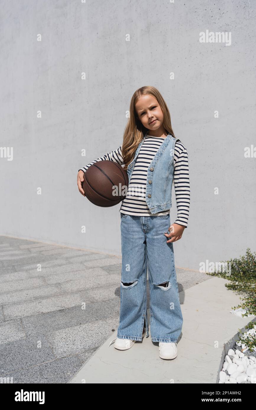 full length of preteen girl in denim vest and blue jeans holding basketball near mall building,stock image Stock Photo