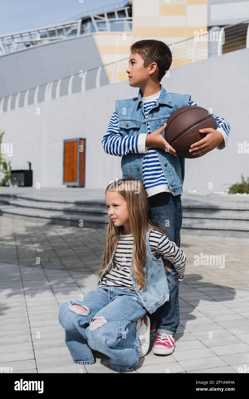girl in stylish clothes hugging legs of boy with basketball standing
