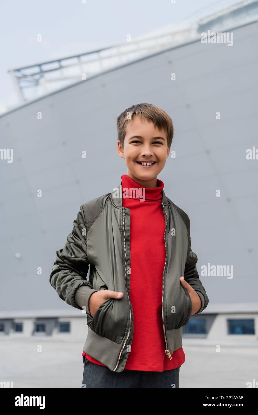 positive preteen boy in stylish bomber jacket and red turtleneck posing with hands in pockets near mall,stock image Stock Photo