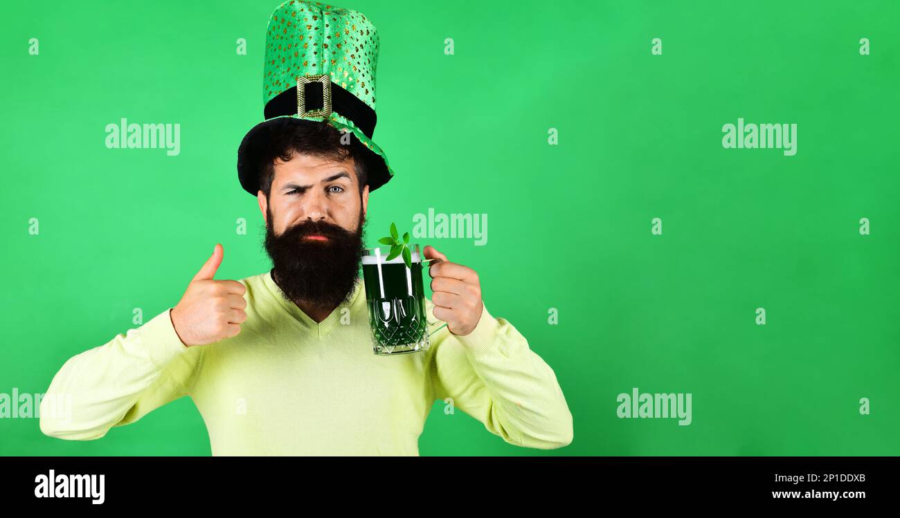 Patricks Day pub party. Serious man with glass green beer shows thumb up. Bearded man holding green beer with shamrock on Patrick day. Handsome male Stock Photo