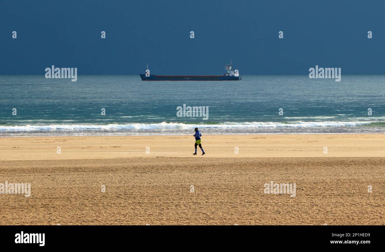 Jogger on a beach on a winter morning with the General Cargo ship HAV Marlin anchored waiting to go into port Sardinero Santander Cantabria Spain Stock Photo
