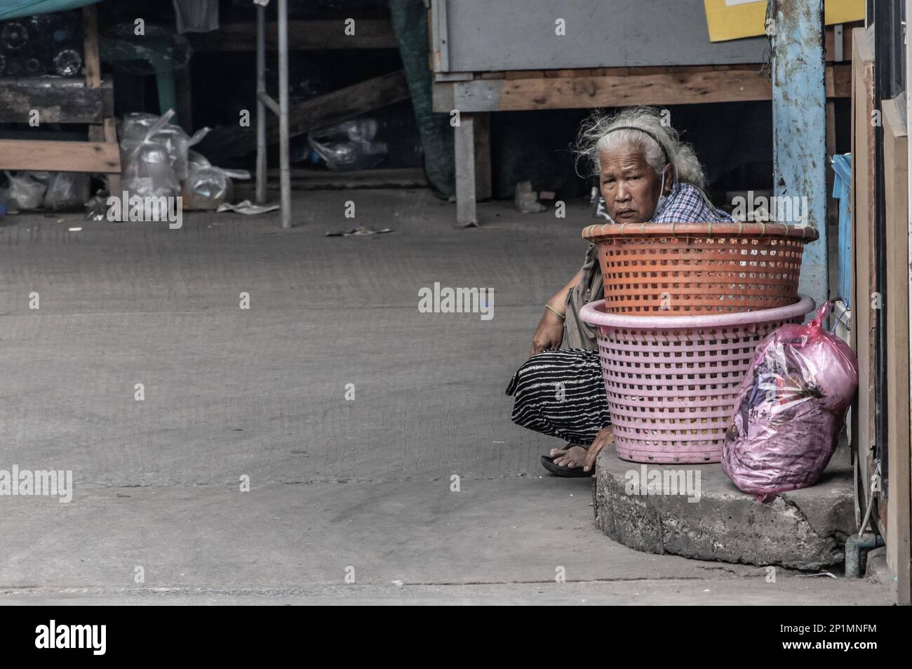 SAMUT PRAKAN, THAILAND, FEB 17 2023,  An old woman sits on the street next to a empty baskets Stock Photo