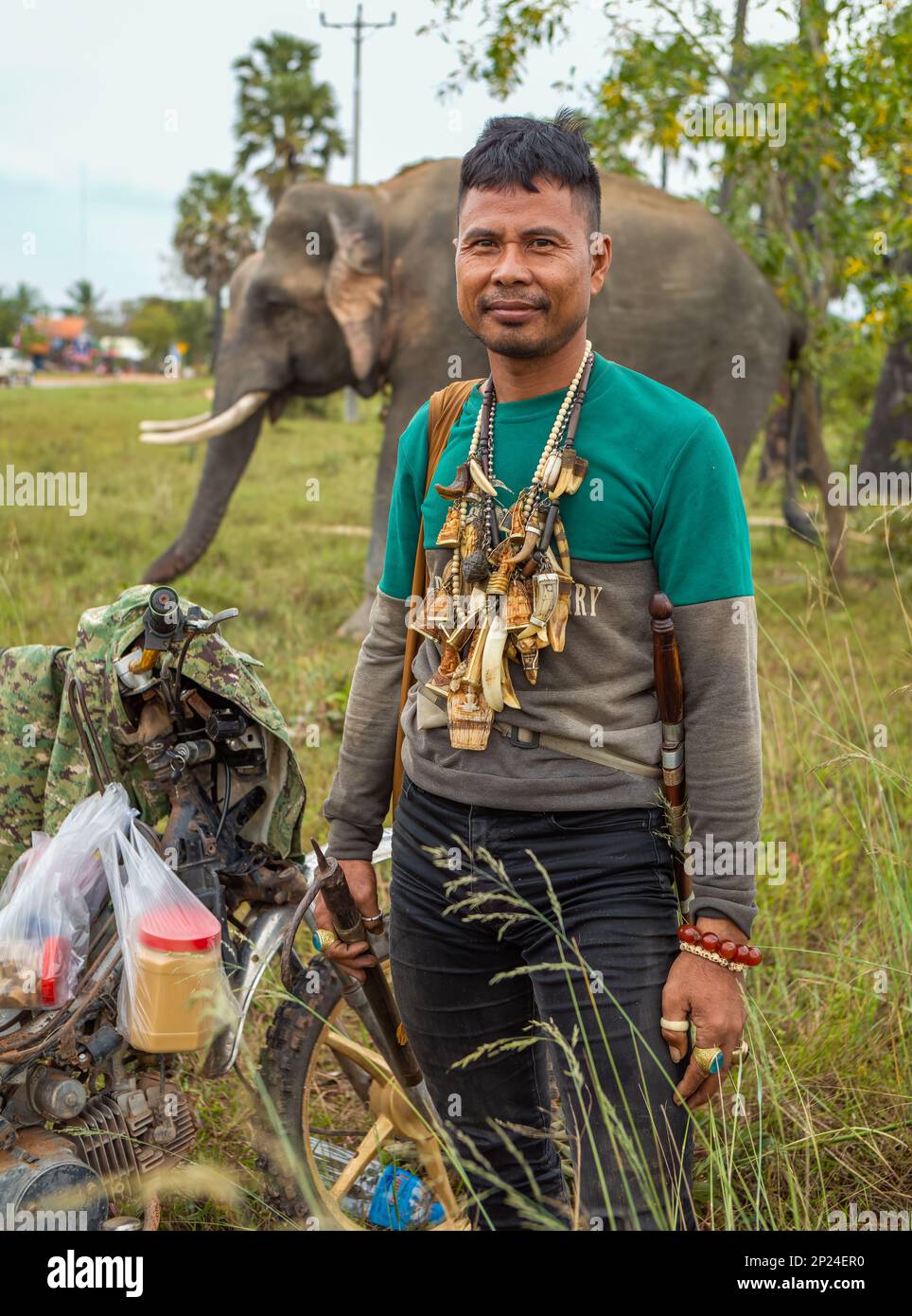 A Cambodian Mahout adorned with Buddhist amulets to ward off danger stands close to his elephant on scrub land in Phumi Khna, Siem Reap Province, Camb Stock Photo