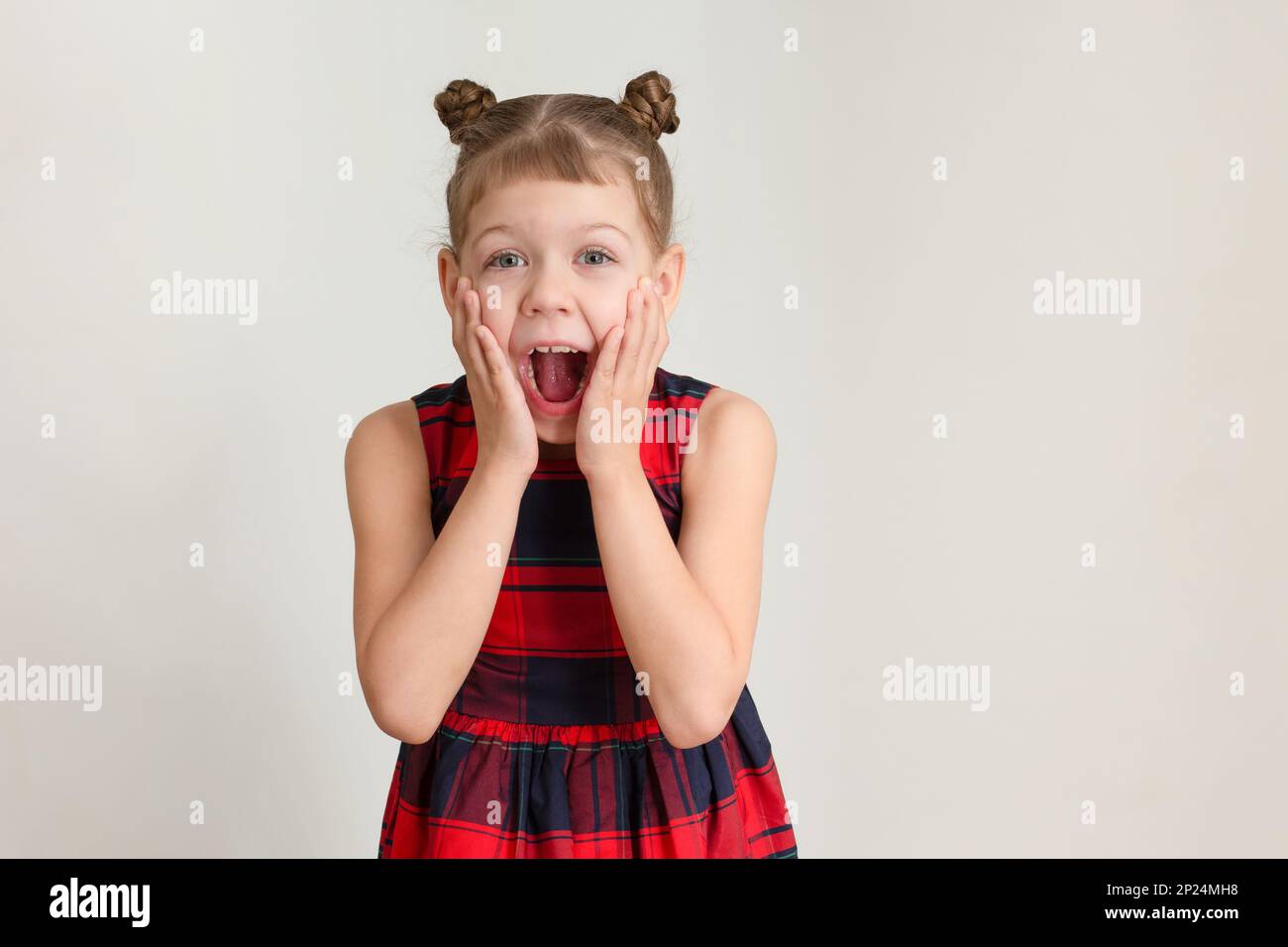 Funny surprised shocked with wide open mouth caucasian child of 6 7 years in red plaid dress holding hands on cheeks because of astonishment Stock Photo