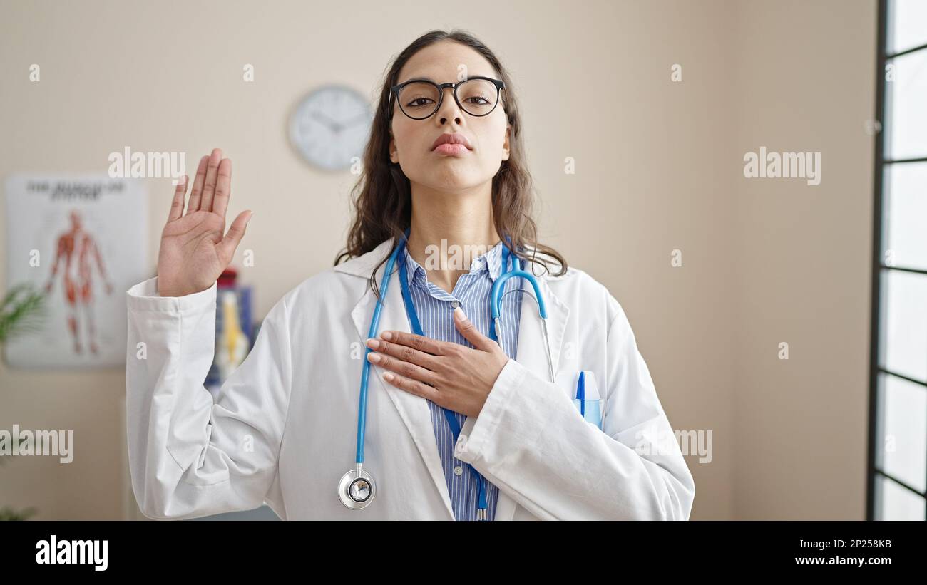 Young beautiful hispanic woman doctor making an oath with hand on chest at clinic Stock Photo