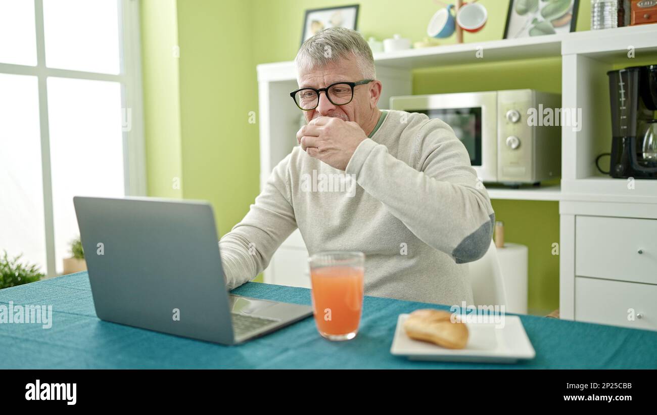 Middle age grey-haired man having breakfast using laptop at home Stock Photo