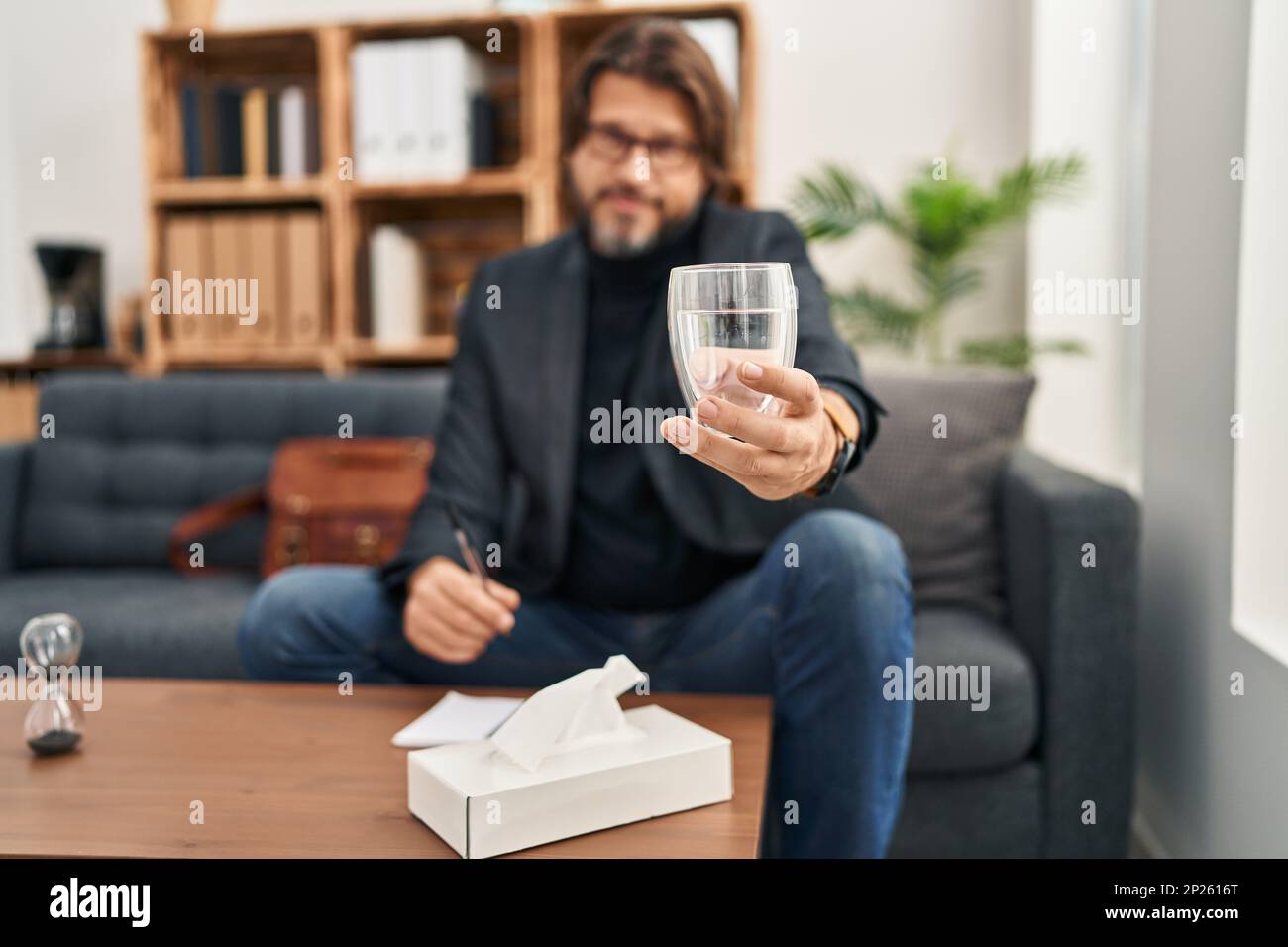 Middle age man psychologist holding glass of water sitting on sofa at