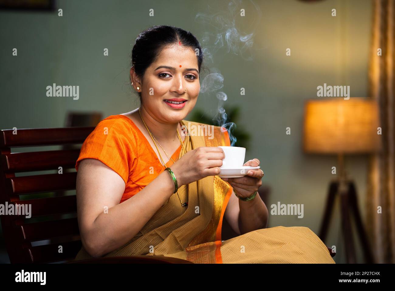 Happy smiling woman drinking tea or coffee by looking at camera while sitting on chair at home - concept of mourning routine, healthy diet and Stock Photo