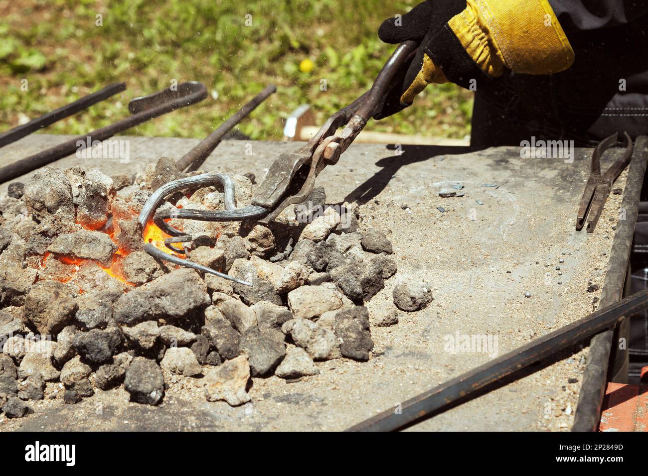 Blacksmith hand holding a metal swirl in tongs on the red hot glowing coals. Forge tools being used in an outdoor smithy Stock Photo
