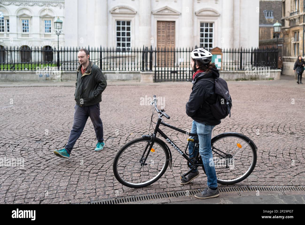 Male cyclist seen taking a rest in the historic University city. A passerby is seen walking from a famous University building in the background. Stock Photo