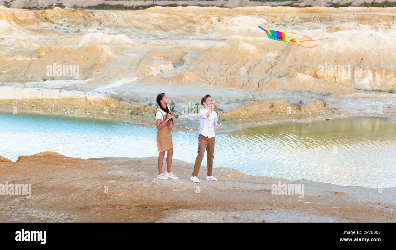 A boy and a girl launch a bright kite standing on the edge mountain near a pond. Stock Photo