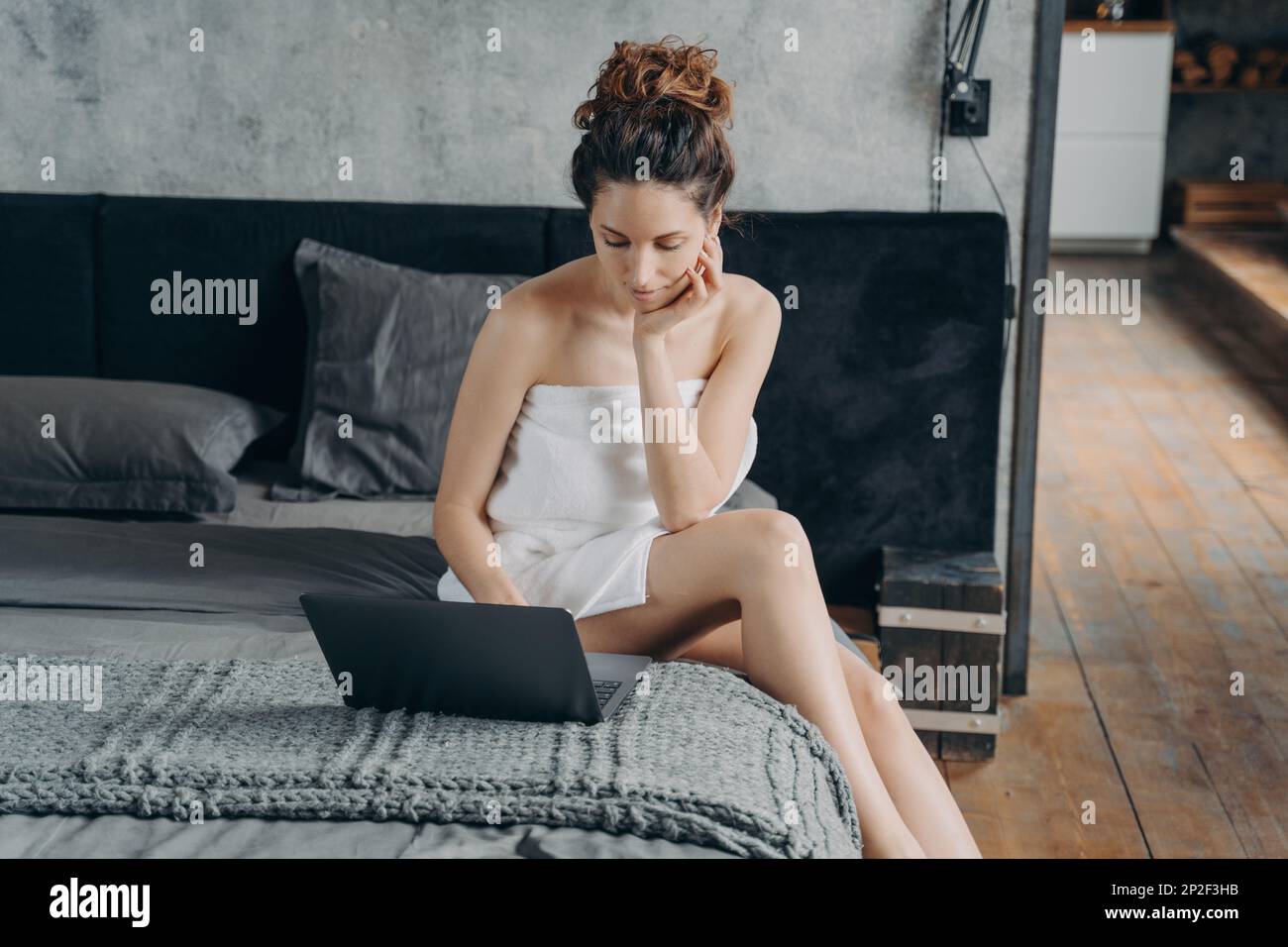 Focused spanish woman wrapped in towel after shower working on laptop sitting on bed. Pensive