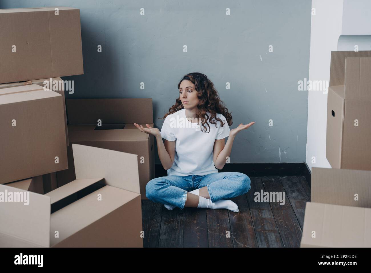 Puzzled young girl shrugging, sitting on the floor with cardboard boxes, having problem with moving to new home. Hard choice, uncertainty, dilemma and Stock Photo