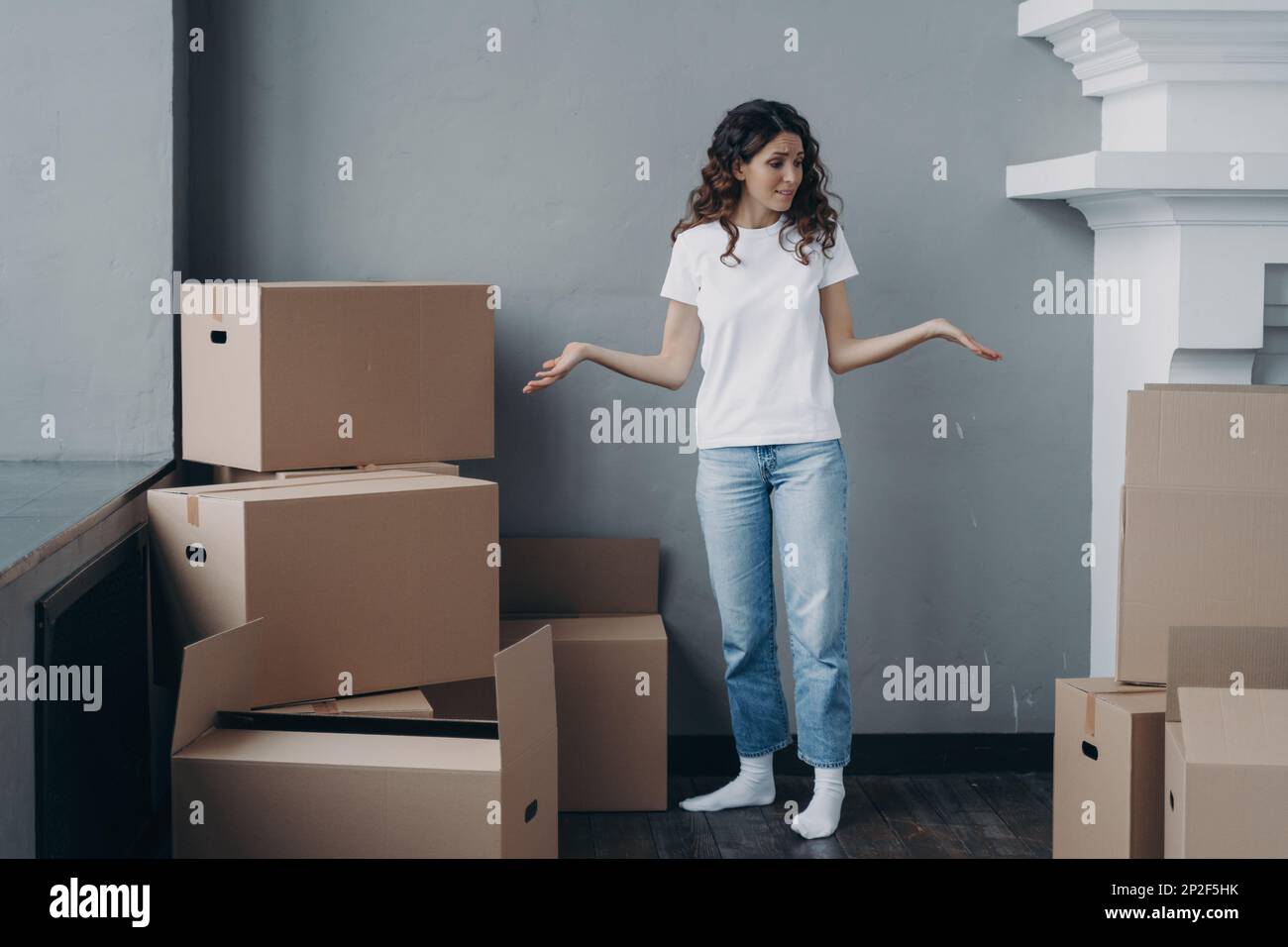 Puzzled female tenant shrugging looking around, suffers stress with packing things and leaving rented apartment. Tired girl standing with cardboard bo Stock Photo