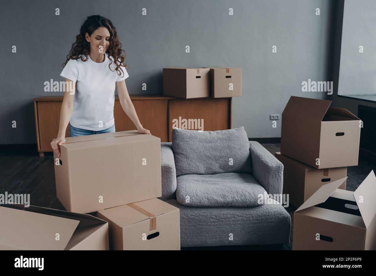 Smiling female tenant with cardboard boxes gathering things for relocation. Pleased hispanic woman renter packing belongings, preparing to move in fir Stock Photo