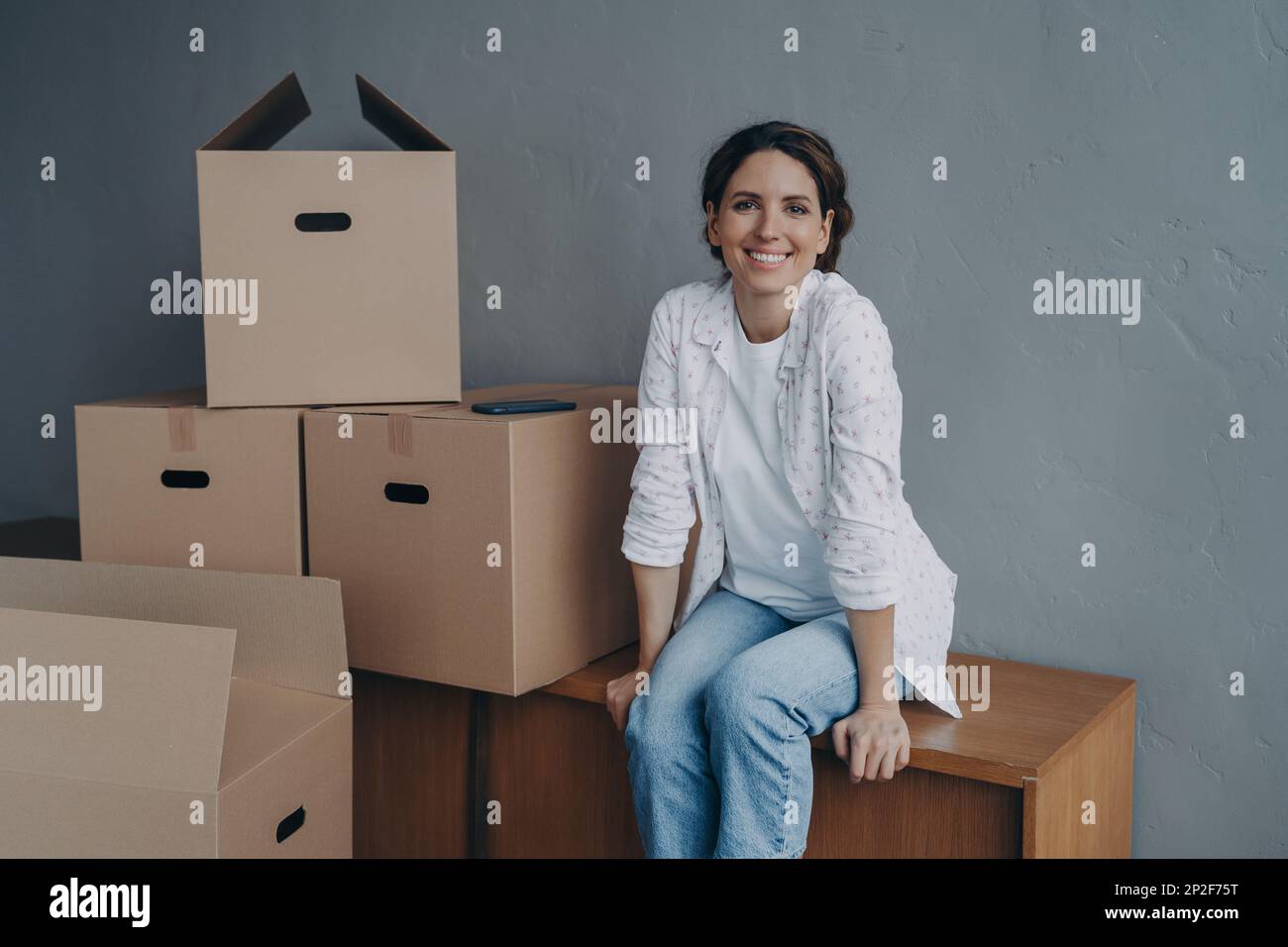 Smiling hispanic woman sitting with cardboard boxes in new rent house or apartment. Mid adult female satisfied with preparation of carton parcels on m Stock Photo