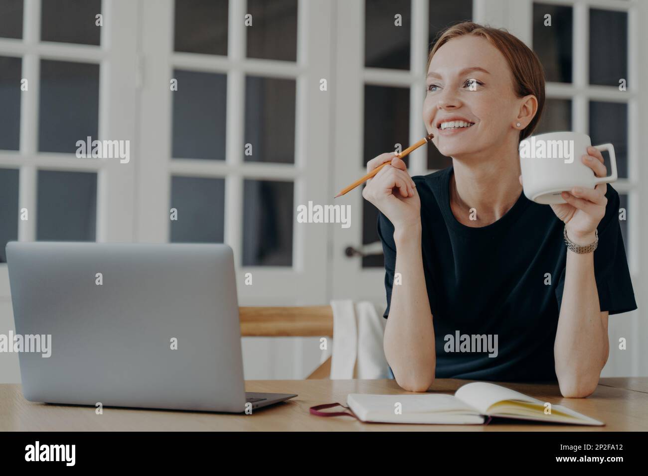 Smiling pensive woman holding pencil and cup write list in notebook, sitting at desk, looking to aside, dreaming, thinking. Happy dreamy female notes Stock Photo