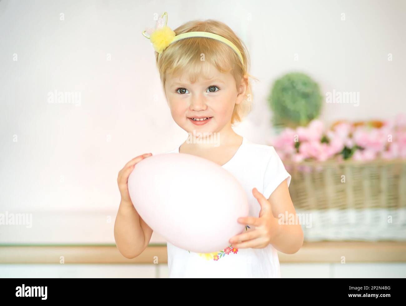 A smiling blonde girl is holding a large pink Easter egg in a bright kitchen with flowers. kid celebrates Easter Stock Photo