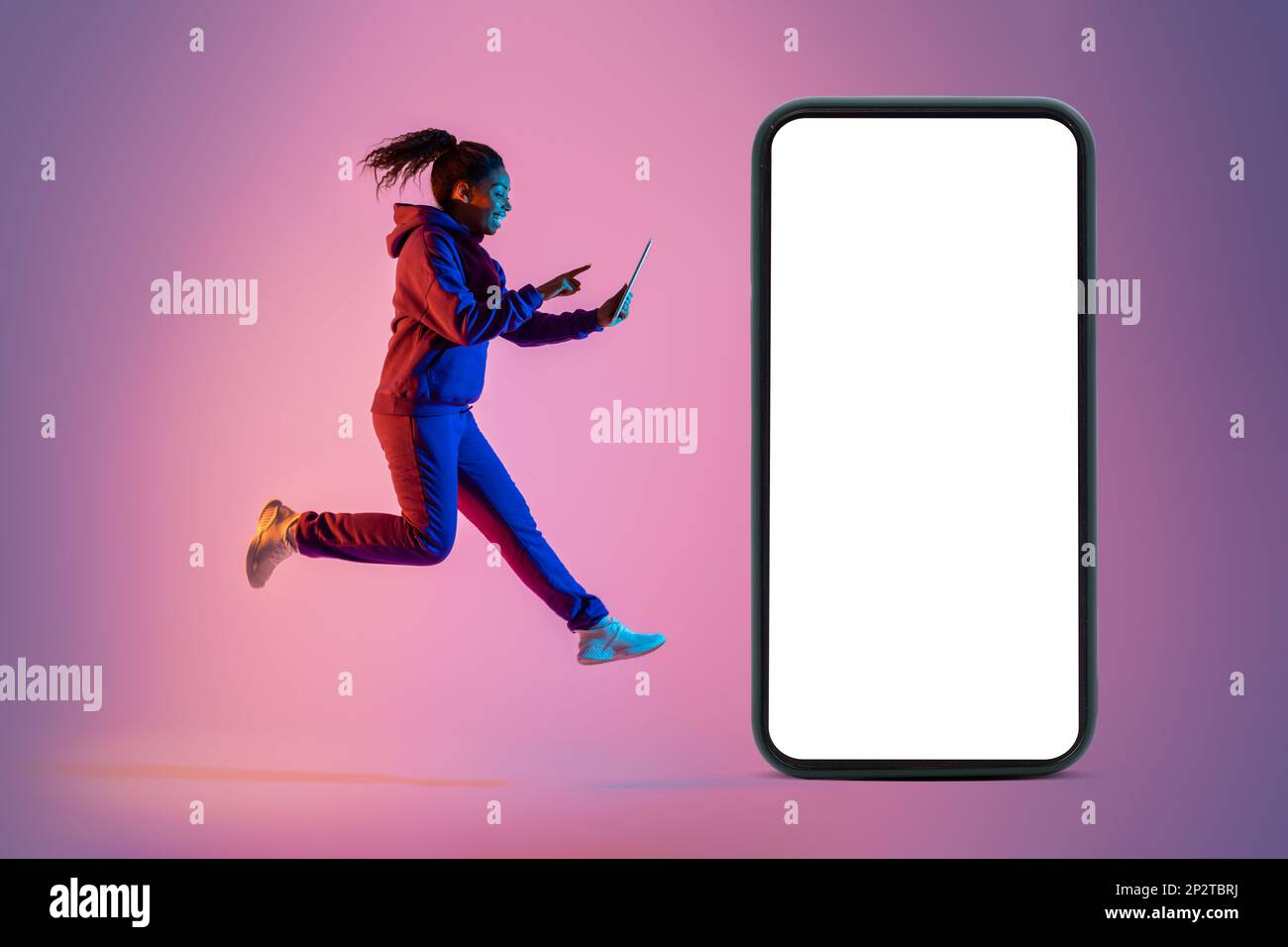 Young black lady jumping with digital tablet next to giant smartphone