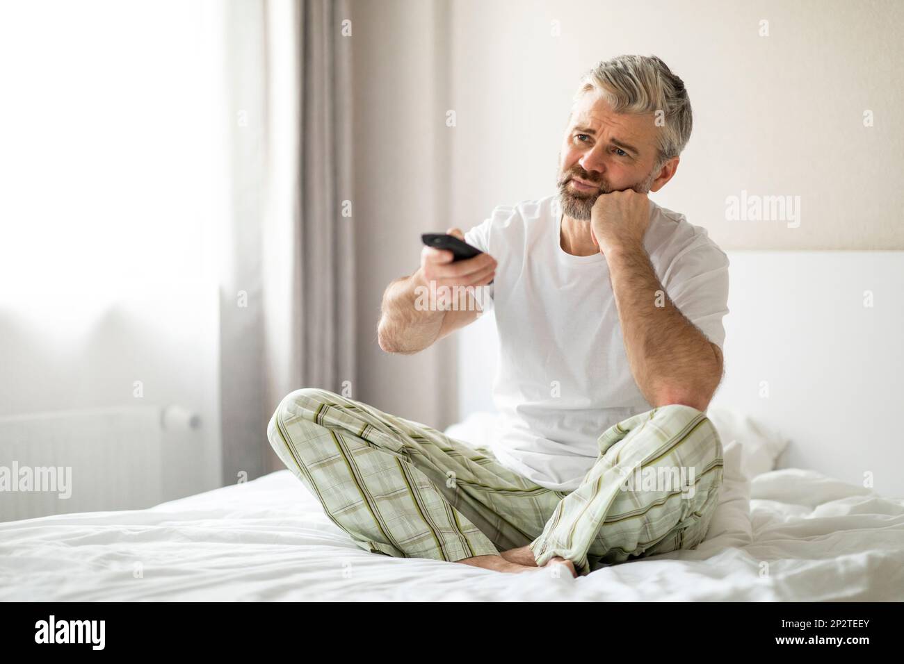 Bored man sitting on bed at home, watching tv Stock Photo