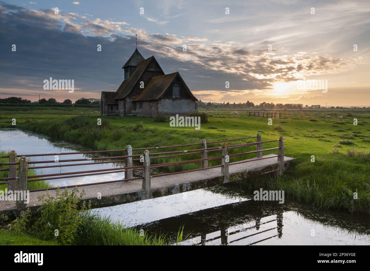 View of grazing moor with footbridge over flooded ditch and church at sunset, St Thomas a Becket Church, Fairfield, Walland Marsh, Romney Marsh Stock Photo