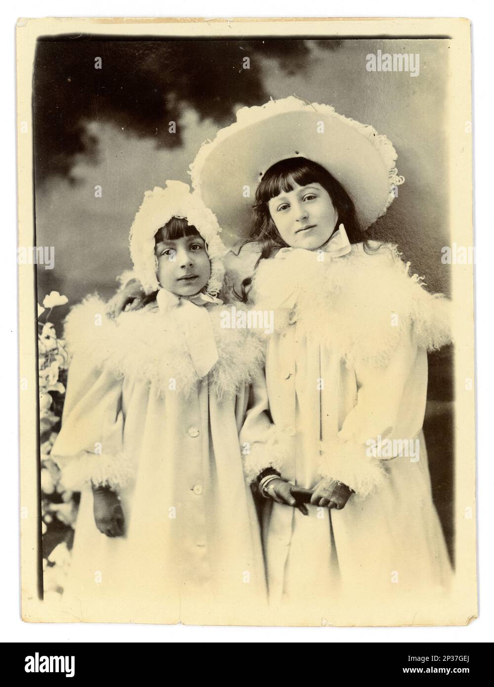 Original Victorian photograph of 2 smiling young  girls, siblings, in white coats trimmed with ostrich feathers, matching bonnet and hat, wearing gloves. Circa 1898, Worcester area, U.K. Stock Photo