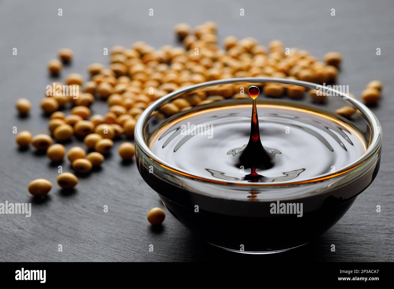 soy sauce with spalsh and drop in glass bowl and dry soybeans on black stone background Stock Photo