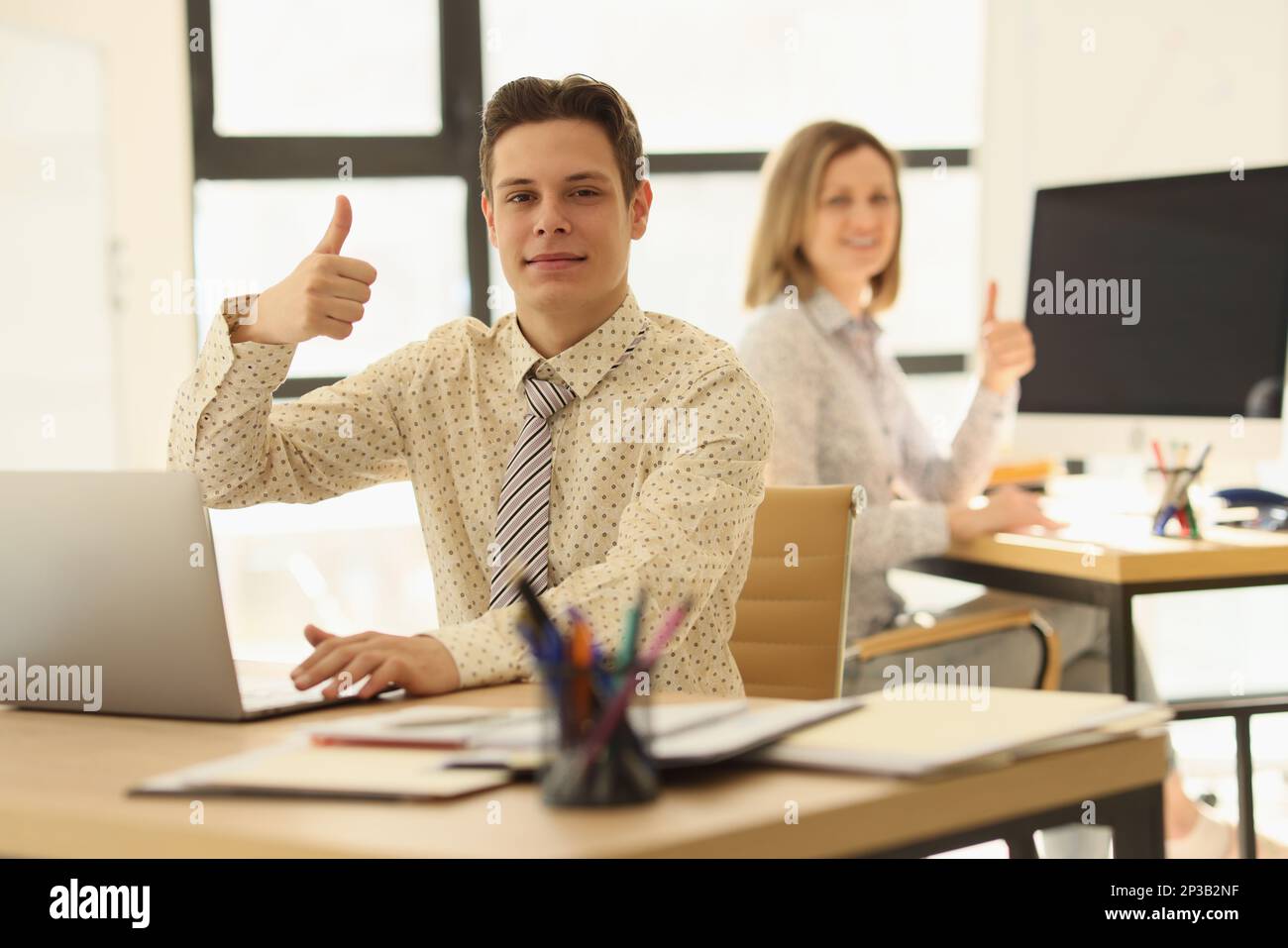 Two managers are sitting at their desks in office and showing thumbs up ...